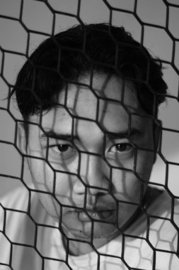 Asyalı bir adam çitin arkasında ifadesiz bir şekilde kameraya bakıyor. Portreli ya da dikey açılı siyah beyaz fotoğraf. Yakın çekim ve vesikalık fotoğraf.