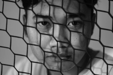 Asyalı bir adam çitin arkasında ifadesiz bir şekilde kameraya bakıyor. Siyah beyaz fotoğraf. Yakın çekim ve vesikalık fotoğraf.