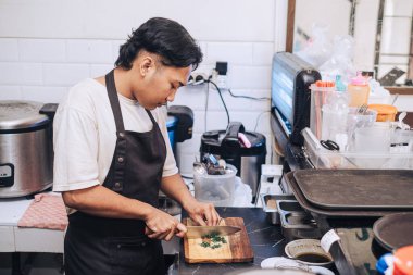 Önlüklü bir Güneydoğu Asyalı şef, öğlenleyin mutfağın tahta kesme tahtasındaki pırasaları kesiyor.