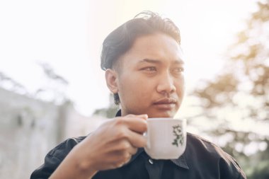 Güneydoğu Asyalı bir adam gün batımında sarhoş olmak için elinde bir fincan kahve tutuyor. Önde ve arkada Bokeh var.