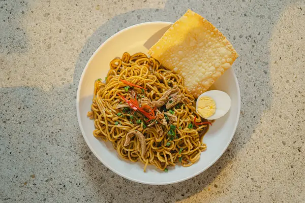 stock image Indonesian Fried Noodles with boiled egg, crackers, chili, and leeks in a white bowl on a stone table as background. Flay lay, overhead, or top view angle.