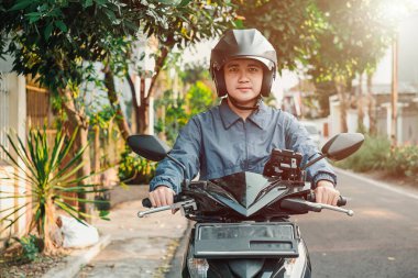 Mavi ceketli Asyalı bir adam otomatik scooter ya da motosiklet kullanıyor, elinde telefon kılıfı var ve öğleden sonra gün batımında kameraya bakıyor. Bokeh veya bulanık arkaplan.