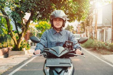 Mavi ceketli Asyalı bir adam otomatik scooter ya da motosiklet kullanıyor, elinde telefon kılıfı var ve öğleden sonra gün batımında kameraya bakıyor. Bokeh veya bulanık arkaplan.