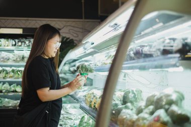 Asyalı bir kadın, Bokeh önplanı ve geçmişi olan bir süpermarketten taze sebze alıyor.