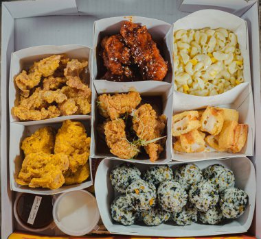 Bir paket servis kutusunda Kore usulü kızarmış tavuk yemekleri var. Kutuda kızarmış tavuk, tatlı ve baharatlı tavuk, makarna ve peynir, kızarmış tofu ve pirinç topları var. Güney Kore 'nin popüler fast food yemekleri.