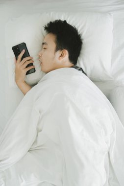 Genç Asyalı adam beyaz yatakta akıllı telefonunu tutarken uykuya dalıyor.