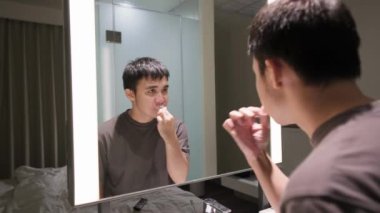 Genç Asyalı adam yatmadan önce modern banyoda aynanın önünde dişlerini fırçalıyor.