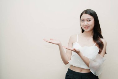 Genç Asyalı kadın, izole edilmiş arka planda gülen yüz ifadesiyle sunum jesti yapıyor. Süslü beyaz bluz ve siyah pantolon giyiyor..