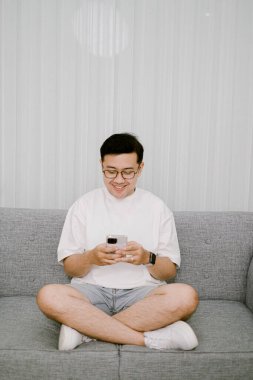 Gülümse, Güneydoğu Asyalı adam, modern bir oturma odasında gri bir koltukta oturmuş, elinde akıllı bir telefonla mutlu bir şekilde sohbet ediyor.