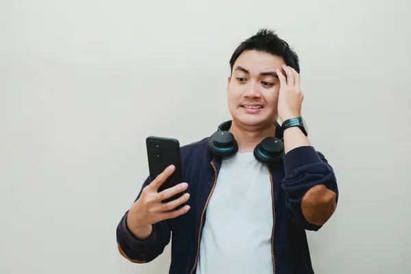 Beyaz tişörtlü ve kulaklıklı mavi kazaklı Asyalı adamın şok olmuş yüzü izole edilmiş arka plandaki akıllı telefona bakarken...