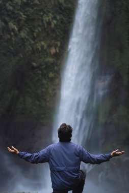 Mavi kapüşonlu bir adam, kolları açık, uzun bir şelaleye bakıyor. Seyahat ve özgürlük.