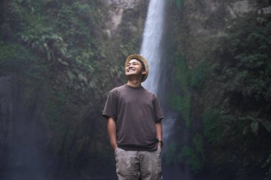 Şelalenin önünde duran, arka planda bokeh olan Asyalı adamın gülen yüzü.