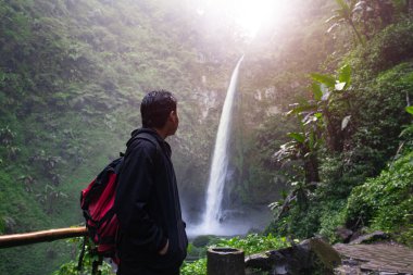 Güneydoğu Asyalı bir adam seyahat ediyor ve güneşin altındaki şelaleye bakıyor.