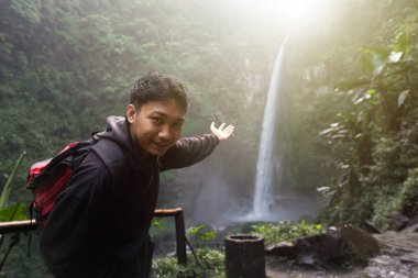 Şelaleyi gösterirken gülümseyen yüz ifadesiyle selfie çeken genç Asyalı adam.