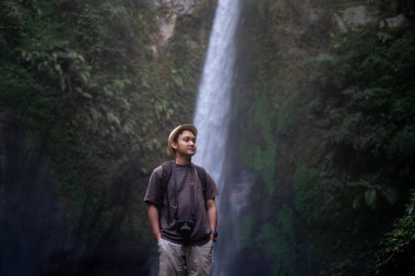Hasır şapkalı ve sırt çantalı bir adam güvenle ayakta duruyor ve şelalenin önünde kolları havada bir kamera taşıyor. Seyahat fotoğrafçılık konsepti.