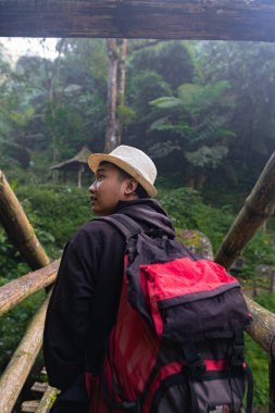 Hasır şapkalı ve sırt çantalı Güneydoğu Asyalı adam Endonezya 'da güneş ışığının altında ahşap bir köprüden geçiyor.