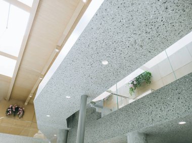 Açık ahşap panelli tavanlı, benekli beton yapılar, cam korkuluklar ve pembe asılı çiçekler de dahil olmak üzere yeşillik içeren çağdaş bir binanın iç mimarisi..