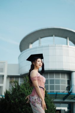 Modern kampüs binasının önünde geleneksel kıyafetlerle mezun olan gururlu Güneydoğu Asyalı kadın.