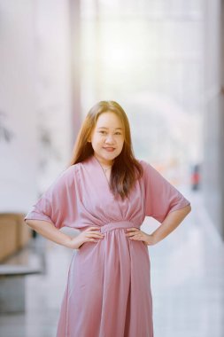 Kendine güvenen Güneydoğu Asyalı kadın pembe elbiseli elleri ile gülümsüyor. Kapalı, parlak, modern, arka planlı bir koridorda duruyor.