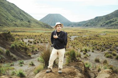 Şapkalı ve sırt çantalı genç bir Güneydoğu Asyalı Bromo Dağı tepesinde duruyor, Doğu Java, Endonezya 'da, elinde bir kamera, keşfetmeye hazır.