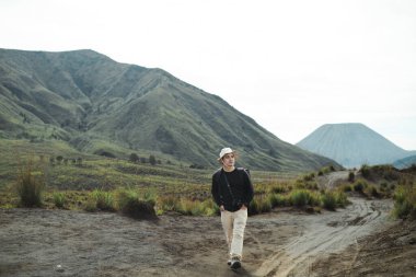 Genç Güneydoğu Asyalı bir adam şapka ve sırt çantası takıyor, Bromo Dağı, Doğu Java, Endonezya 'da kirli bir yolda tek başına yürüyor.
