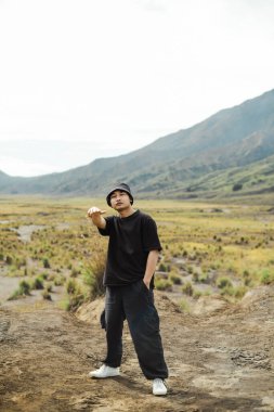 Siyah kıyafetli ve kova şapkalı genç bir Güneydoğu Asyalı Bromo Dağı, Doğu Java, Endonezya 'da kuru çayırlarda duruyor. Arka planda görkemli dağları işaret ederek macera ve keşif ruhunu simgeliyor..