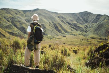 Güneydoğu Asya 'lı genç bir adamın Bromo çimenli tepelerde tek başına yürüyüşünü, bir sırt çantası ve ceket taşımasını, Doğu Java, Endonezya' daki yeşil manzarayla çevrili bir yürüyüş yolunda.