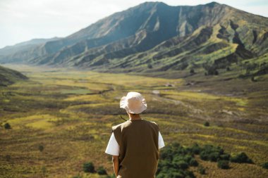 Yeşil volkanik dağlara ve Endonezya, Bromo, Doğu Java 'daki geniş bir vadiye hayran olan, şapka ve yelek giymiş Güneydoğu Asyalı bir erkek yürüyüşçünün arka görüntüsü.