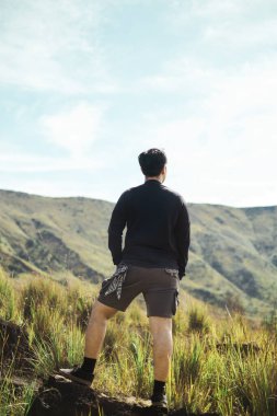 Arkadan görülen genç bir Güneydoğu Asyalı, çimenli bir tepede duruyor, Doğu Java, Endonezya 'daki engebeli engebeli dağ manzarasına derin derin bakıyor.