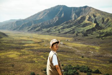 Beyaz gömleğin üzerine bej şapka, gözlük ve kahverengi yelek giyen Güneydoğu Asyalı genç bir adam Bromo vadisinde duruyor ve kameraya doğru hafifçe gülümsüyor..