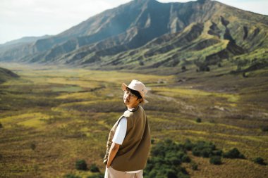 Kovalı şapka ve yelek giyen Güneydoğu Asyalı bir adam Bromo, Doğu Java, Endonezya 'nın arka planında dağlarla dolu çimenli bir vadide duruyor ve gündüz kamerasına doğru bakıyor.. 