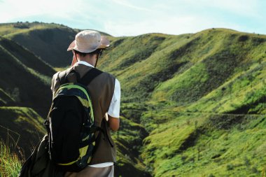 Bromo, Endonezya 'da yeşil volkanik tepelerin keyfini çıkaran, kova şapkalı ve sırt çantalı Güneydoğu Asyalı bir yürüyüşçünün arka görüntüsü.