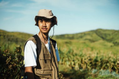 Kovalı şapka ve yelek giymiş genç bir Güneydoğu Asyalı, güneşli yeşil tepelerde ve çimenli arazilerde macera, özgürlük, seyahat ve doğa temaları için mükemmel dururken, mesafeye bakıyor..