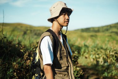 Kovalı şapka ve yelek giymiş genç bir Güneydoğu Asyalı, güneşli yeşil tepelerde ve çimenli arazilerde macera, özgürlük, seyahat ve doğa temaları için mükemmel dururken, mesafeye bakıyor..