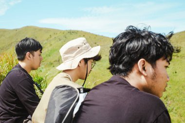 Üç Güneydoğu Asyalı adam, Endonezya 'nın Bromo dağlarındaki parlak güneş ışığı altında canlı yeşil tepelerde yürüyüş yaparken barışçıl bir mola verdiler.