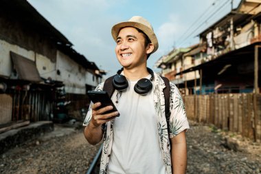 Telefonları ve kulaklıkları olan mutlu bir Güneydoğu Asyalı tren raylarının yakınındaki bir köy sokağında yürüyor. Seyahat, yaşam tarzı ve Güneydoğu Asya keşif temaları için harika.