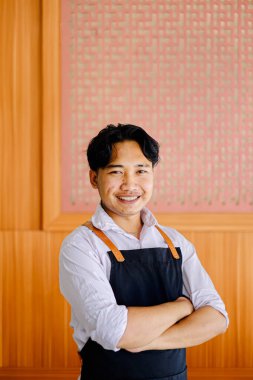 Siyah önlük ve beyaz gömlek giyen gülümseyen Asyalı bir aşçının portresi modern bir ahşap restoranın içinde kolları çapraz duran mutfak, konukseverlik ve iş teması için ideal..