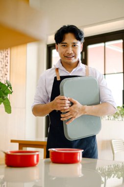 Siyah önlüklü, kendine güvenen genç Asyalı bir adam modern bir restoran mutfağında, profesyonel konukseverlik hizmeti ve verimli yemek hazırlama hizmeti sunan bir tepsi tutuyor.