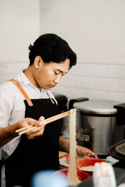 Odaklanmış, siyah önlüklü genç bir Güneydoğu Asyalı şef, yemek çubuklarını, hareketli, modern bir restoran mutfağında sıcak erişte hazırlamak için ustalıkla kullanıyor.