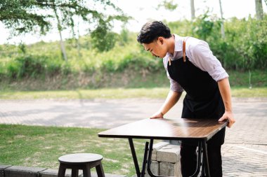 Önlüklü çalışkan genç bir Güneydoğu Asyalı garson açık hava yemek alanında ahşap bir masa hazırlıyor. Restoranı servis için hazırlıyor..