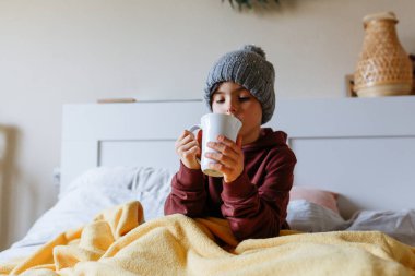 Girl wearing grey warm hat sits on the bed under warm blanket and drinks hot tea clipart