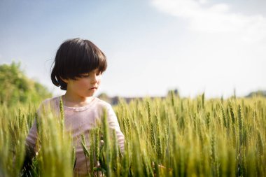 side view of girl with dark short hair in pink top standing in greenwheat field