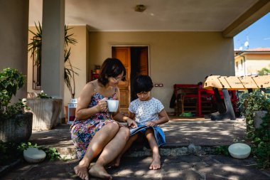 mother and daughter sitting in front of the house watching magazine and drinking coffee