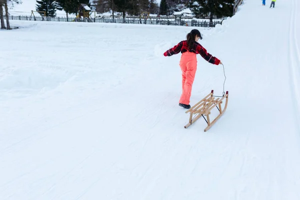 穿着红色套头衫和橙色滑雪裤 一头乌黑的长发 背着木制雪橇在雪地的路上爬山的女孩 — 图库照片