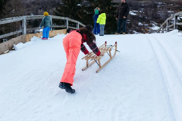 身穿红色套头毛衣和橙色滑雪裤的女孩推着木制雪橇爬上雪山 而其他孩子和家长则在山顶等着 — 图库照片