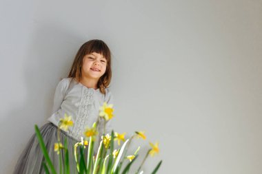 smiling girl with fringe in white top and grey skirt stands behind daffodil flowers