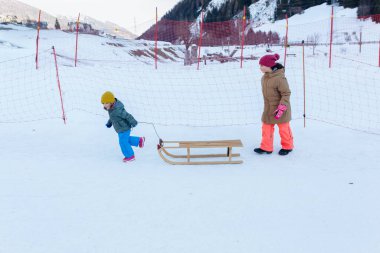 İki çocuk parlak kış elbiseleriyle kar yamacında ahşap kızağı çekiyor.