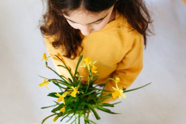 high angle view on child with dark long hair in yellow top holding Easter pot of daffodils