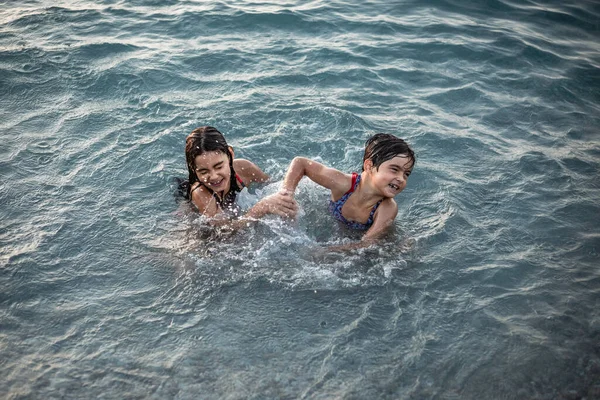 Mavi denizde oynayan, koyu saçlı, gülümseyen iki kızın üst görünüşü.