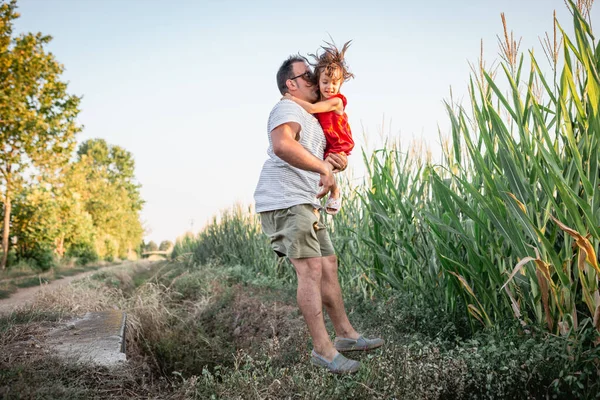 father in striped tee shirt and small daughter in arms jumping over pit in corn field